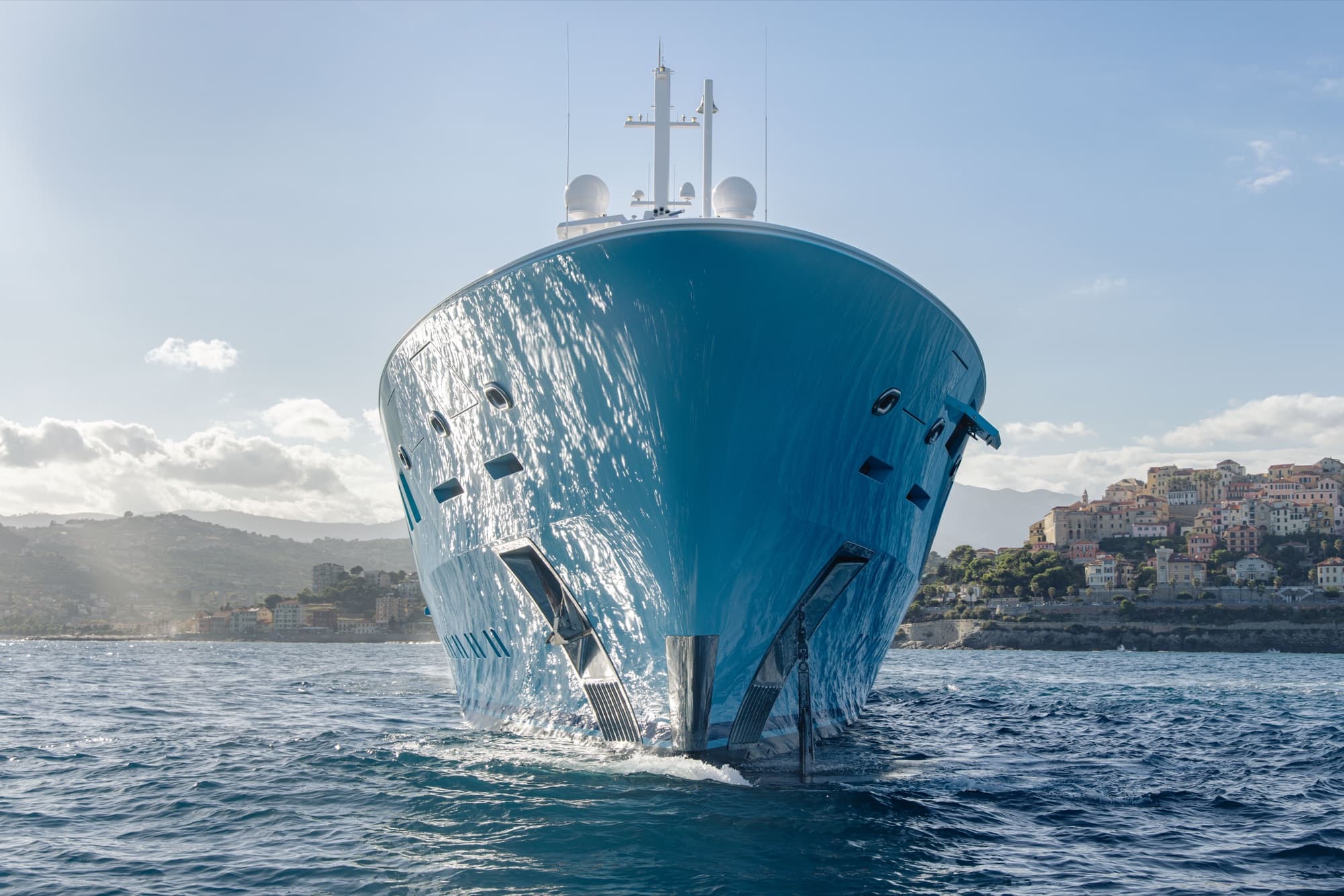 AXIOMA bow-on view at anchor showing the iconic turquoise hull against a Mediterranean coastal village
